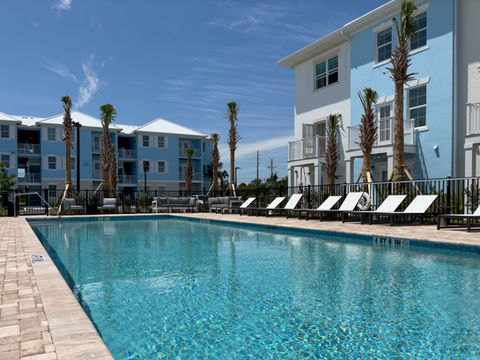 A pool in a residential complex with white lounge chairs, young palm trees, and pastel-colored buildings in the background.