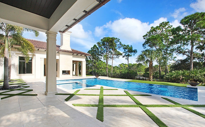 Spacious backyard pool at a luxury residence with geometric grass inlays on the patio, a fire feature, and lush trees in the background under a clear sky.