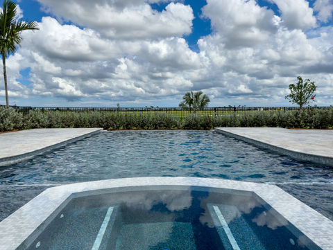 A luxurious pool with a raised spa, surrounded by a wide stone deck and manicured hedges, with an expansive view of the sky and landscape.
