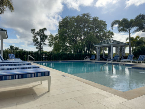 Tranquil pool with a classical gazebo and lounge chairs, encased by lush greenery and a clear, partly cloudy sky above.