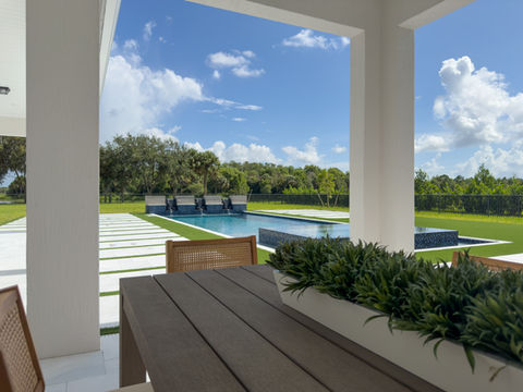 A view from a shaded patio dining area overlooking a modern pool with water features, surrounded by artificial turf and white stone pavers.