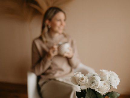 Eine Frau (Désirée Tobler) sitzt unscharf im Hintergrund auf einem weißen Stuhl und hält eine Tasse in den Händen. Im Vordergrund steht ein Glas mit weißen Rosen auf einem Tisch, daneben liegen ein Notizbuch und ein Stift.