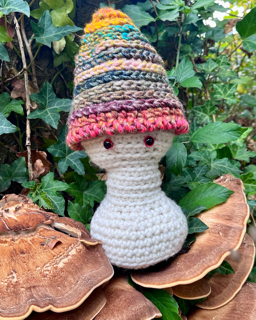 Handmade crochet Mushroom Wisp Morel wearing his Firelight Forest cap beside woodland mushrooms