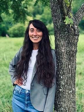 Image of a woman with a light gray jacket and white shirt posing by a tree in a field