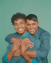 Two influencers standing close together against a teal backdrop, smiling warmly as one person wraps their arms around the other in a friendly embrace.