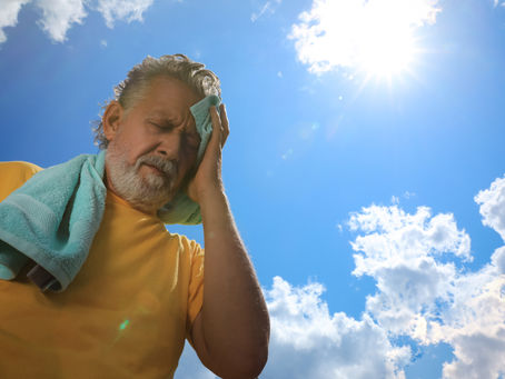 Older man in a yellow shirt wipes his sweaty face with a towel under a bright sun and blue sky, looking overheated.