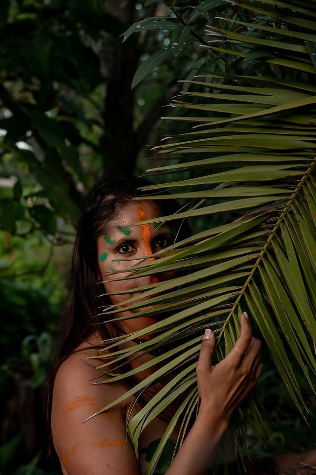 A woman with paint on her face peeks through large palm leaves in a lush, green environment during a brand photoshoot abroad.
