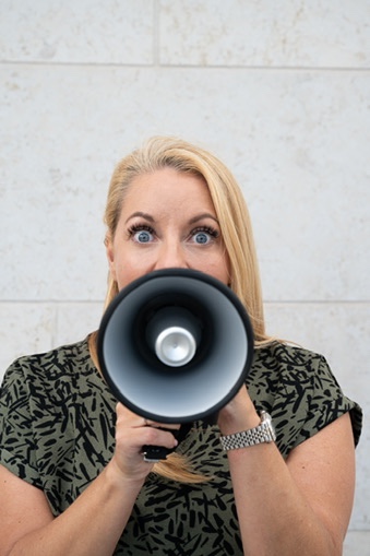 Person with long hair and a megaphone, capturing an element of surprise, embodies the essence of brand photography against a plain wall backdrop.