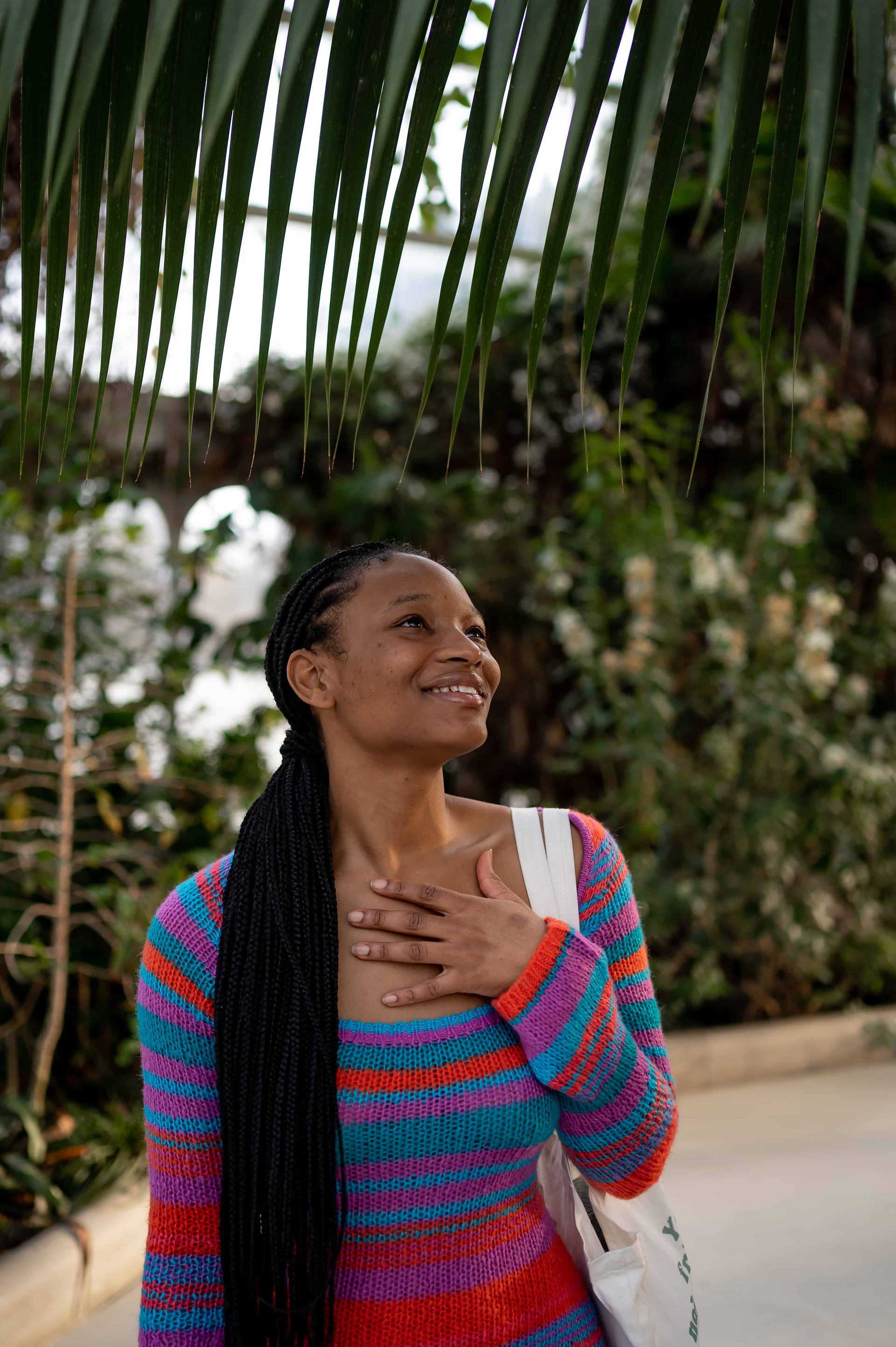 Person wearing a colorful striped sweater smiles while looking up, standing under palm leaves in an outdoor setting.