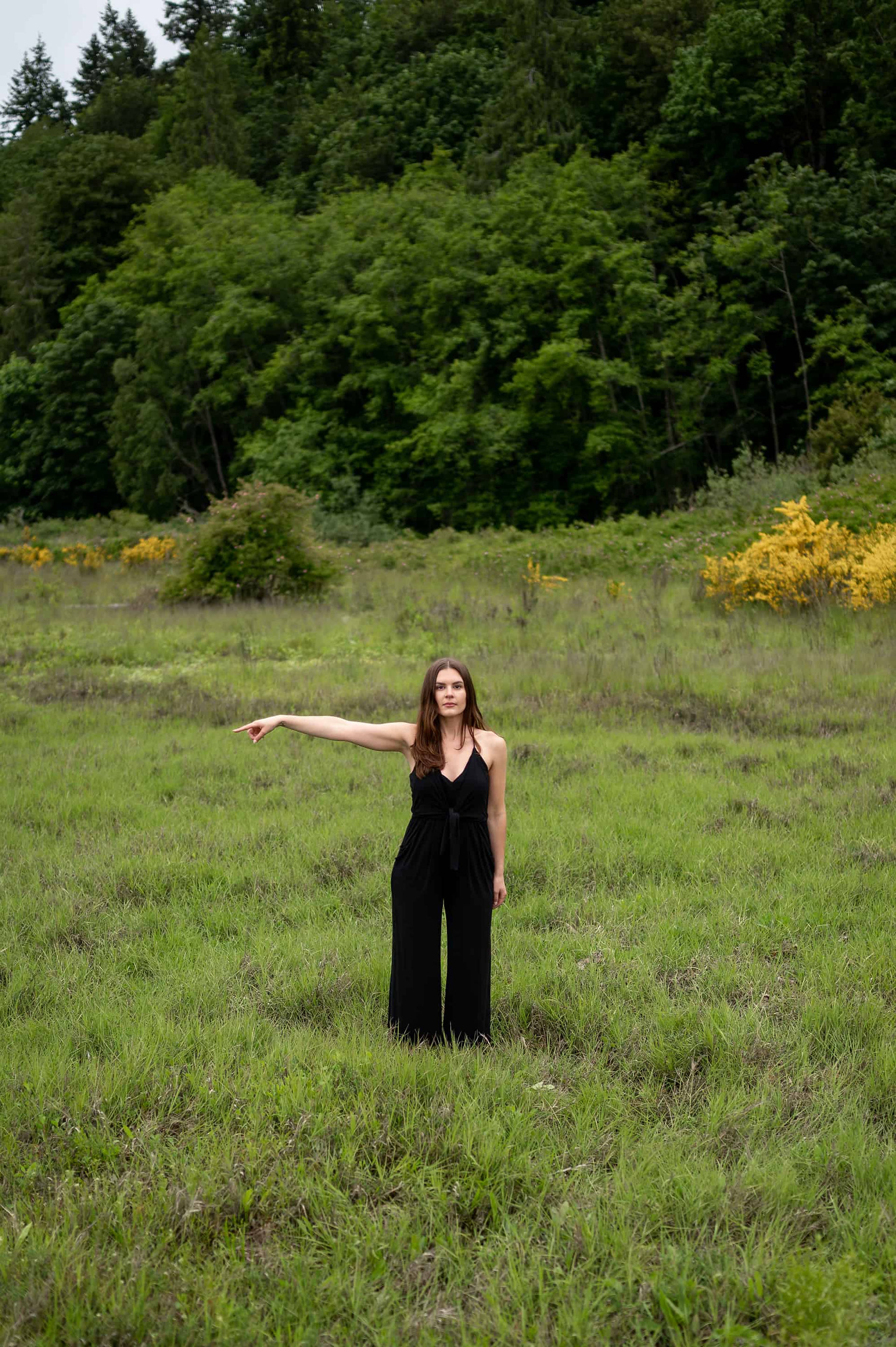 A woman in a black outfit stands in a grassy field with dense trees in the background, pointing with her right arm extended, captured by a personal brand photographer.