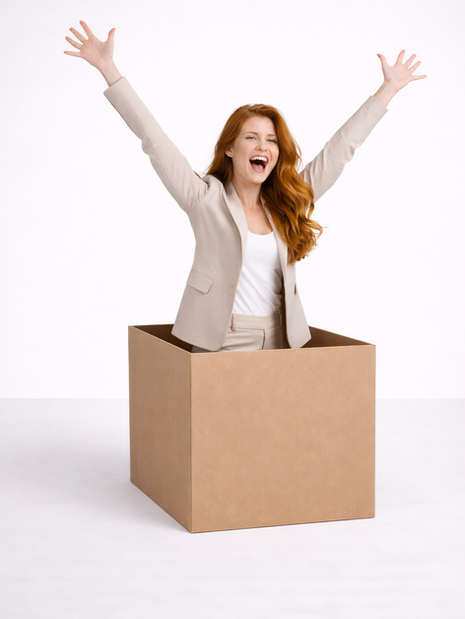 A red-haired woman joyfully throws her arms up while standing inside a cardboard box.