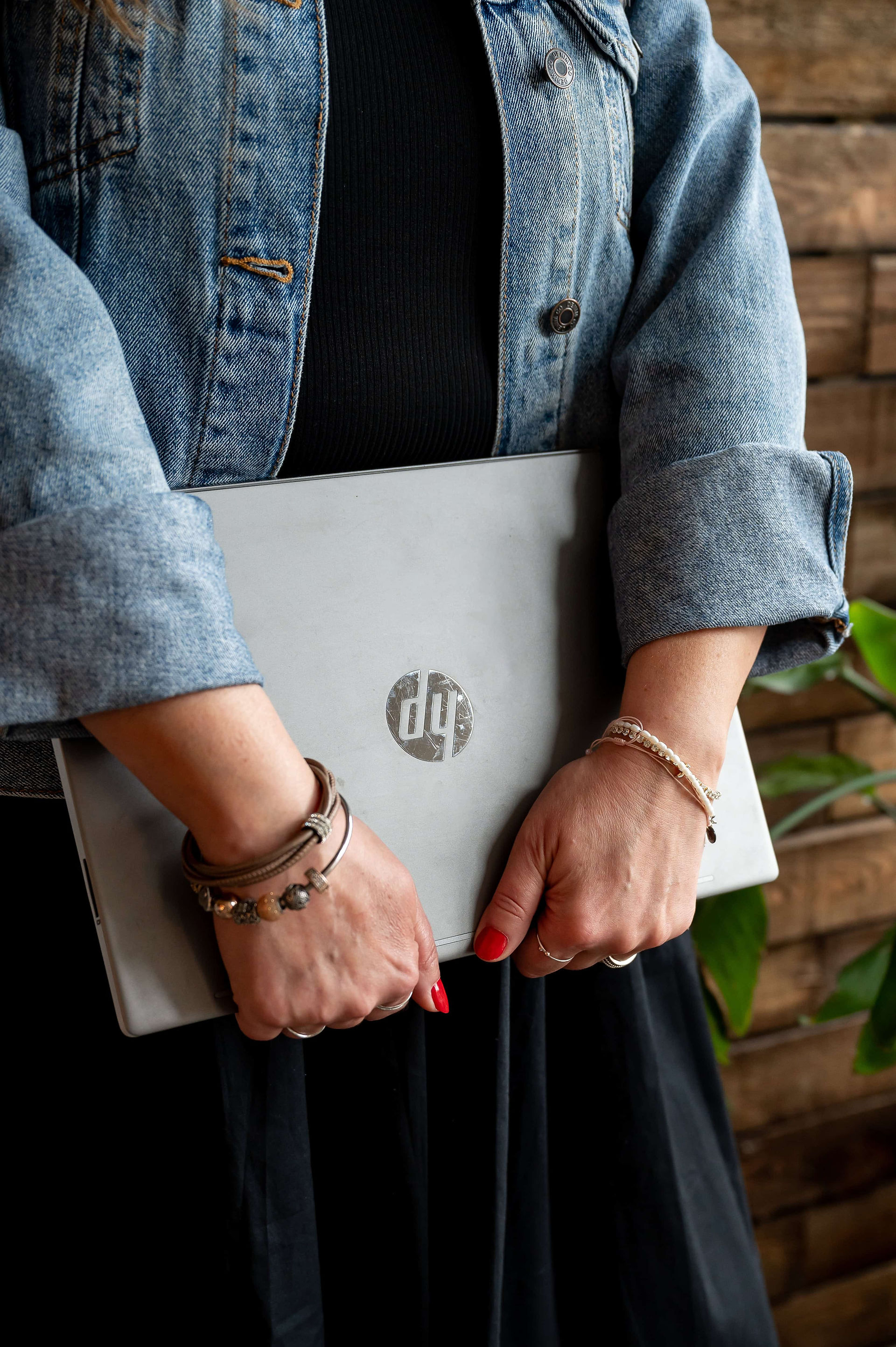 Close up of woman's arms in jean jacket holding a hp laptop upside down. She has red nails and is wearing bracelets.