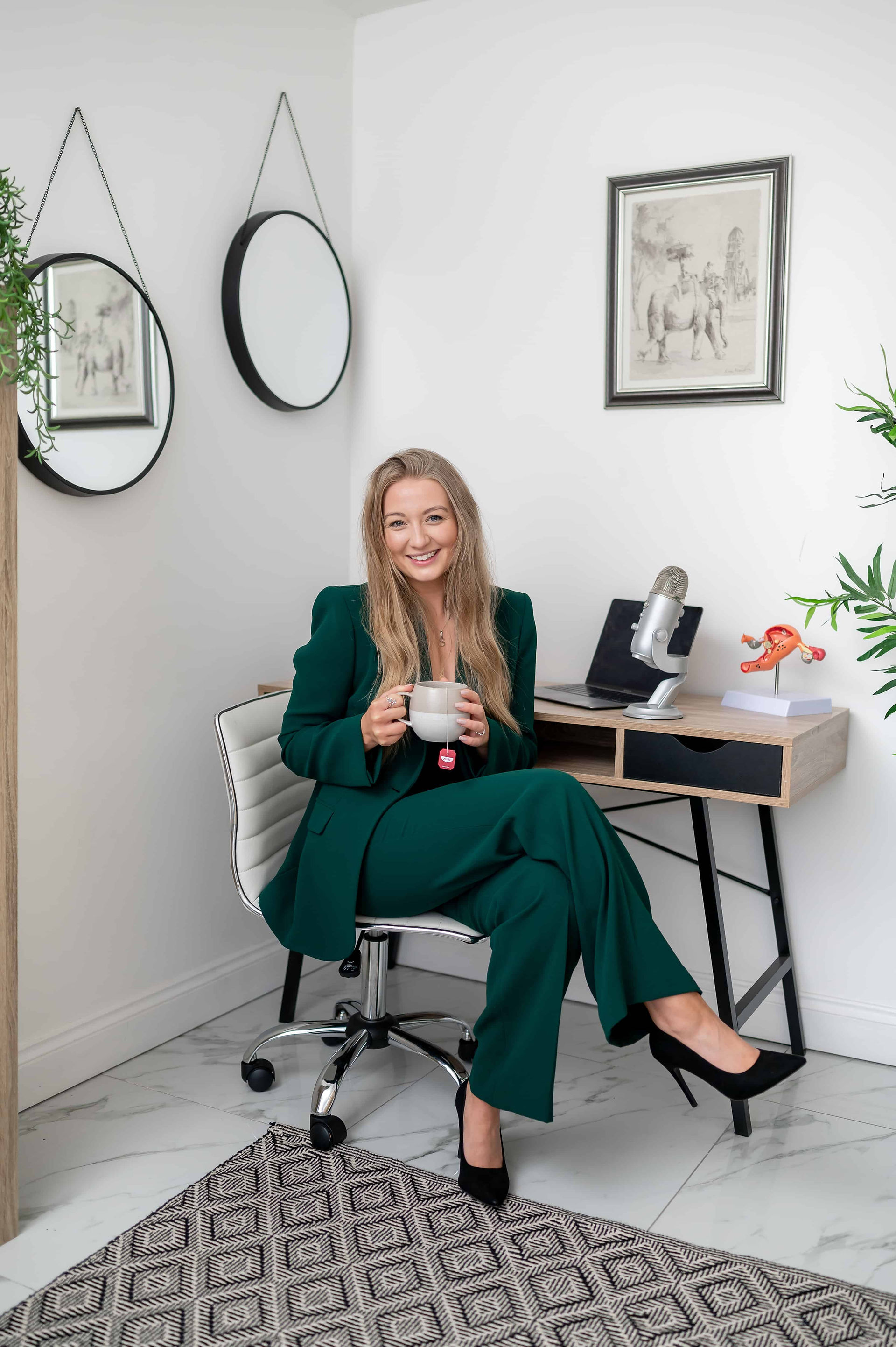 Health coach sitting at office desk in green suit holding cup of pukka tea.