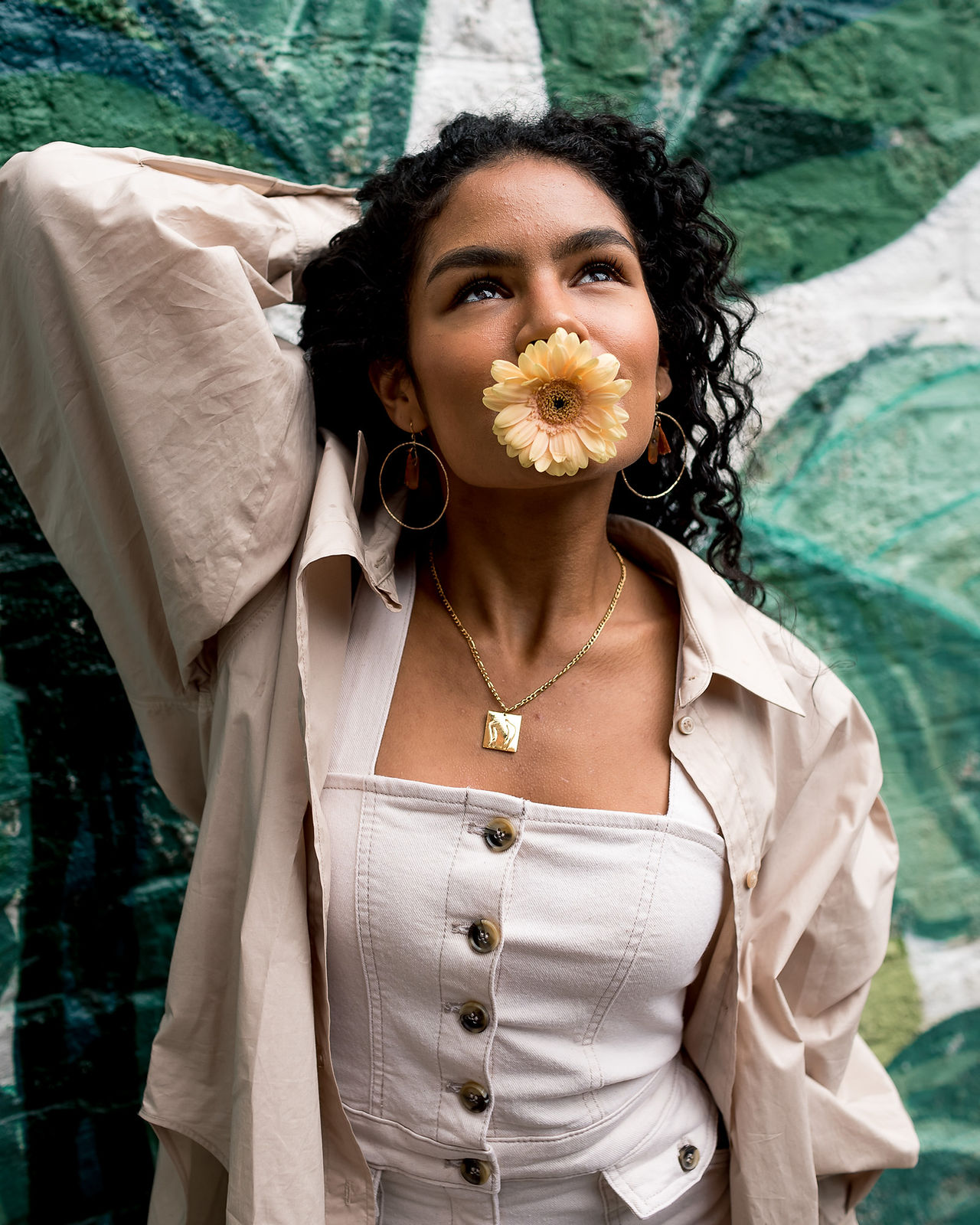 young woman against green painted leafy wall with flower in mouth