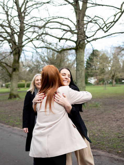 Two people are embracing on a scenic park pathway, framed by lush trees, while another person stands nearby, smiling warmly. It's a perfect moment captured through brand photography.
