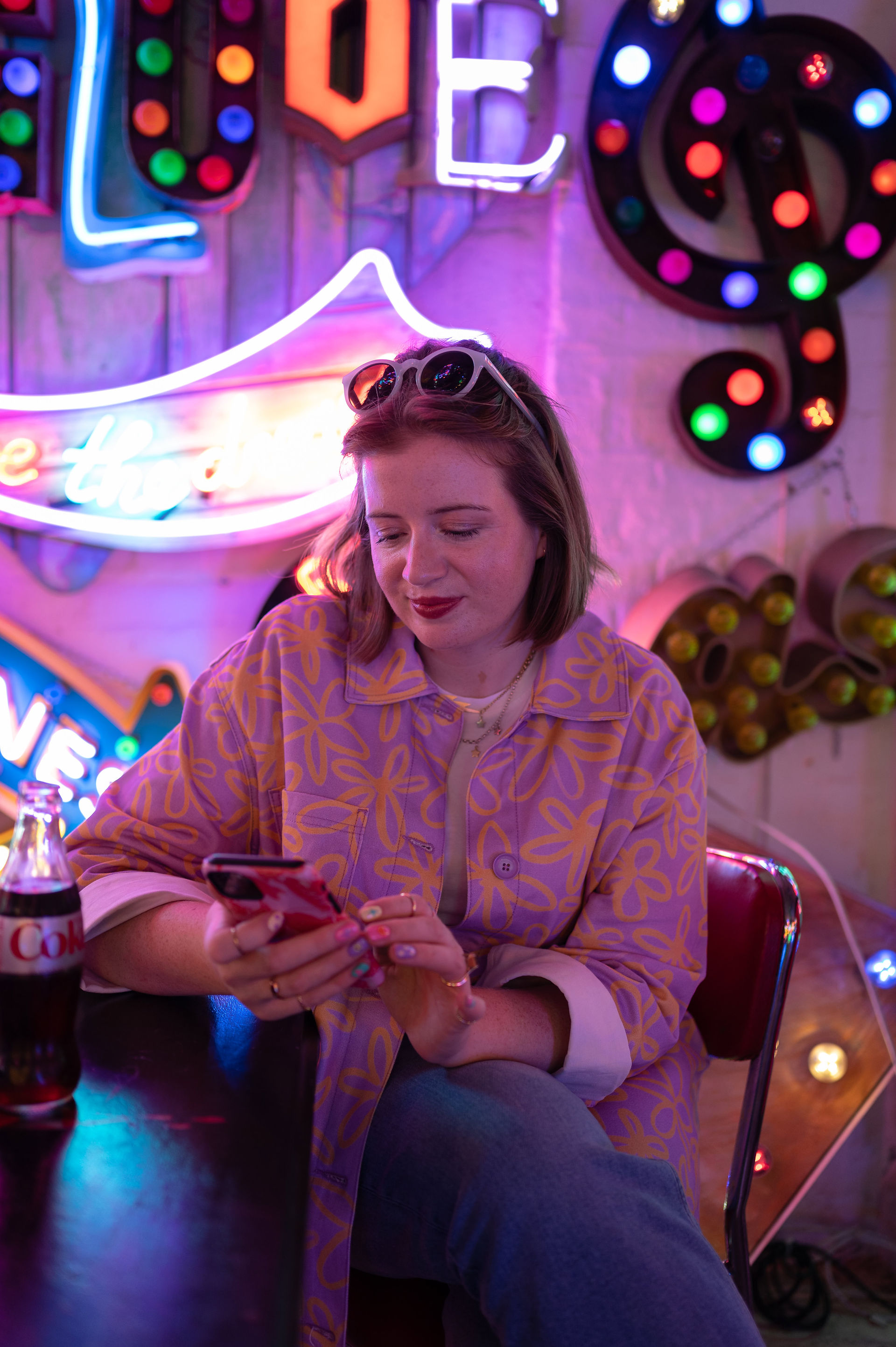 Woman seated indoors, looking at her phone. She wears a patterned shirt and sunglasses on her head. Background features colorful neon signs. A Diet Coke bottle rests on the table.