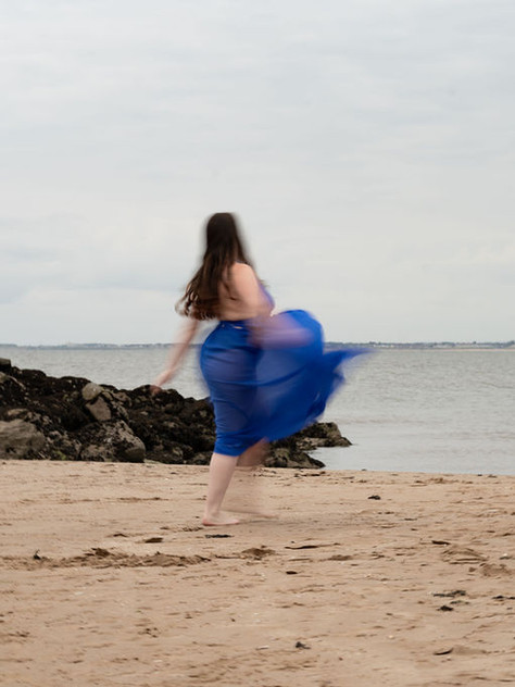 A person in a flowing blue dress spins on a sandy beach near rocky outcrops, with a cloudy sky and calm sea in the background.