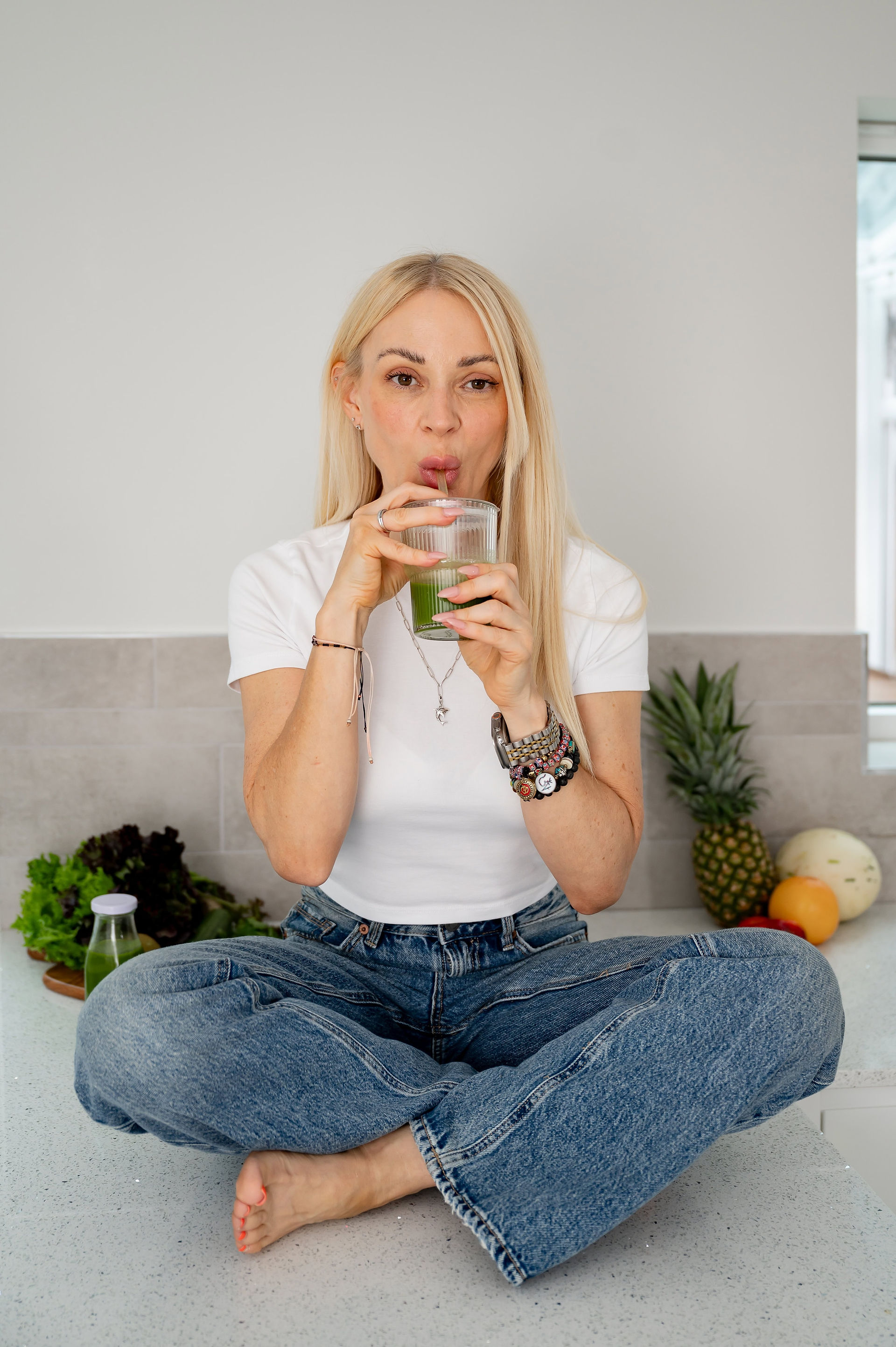 Holistically Balanced Blonde sipping a green smoothie sitting on counter surrounded by fruit during her North Wales brand photography session.