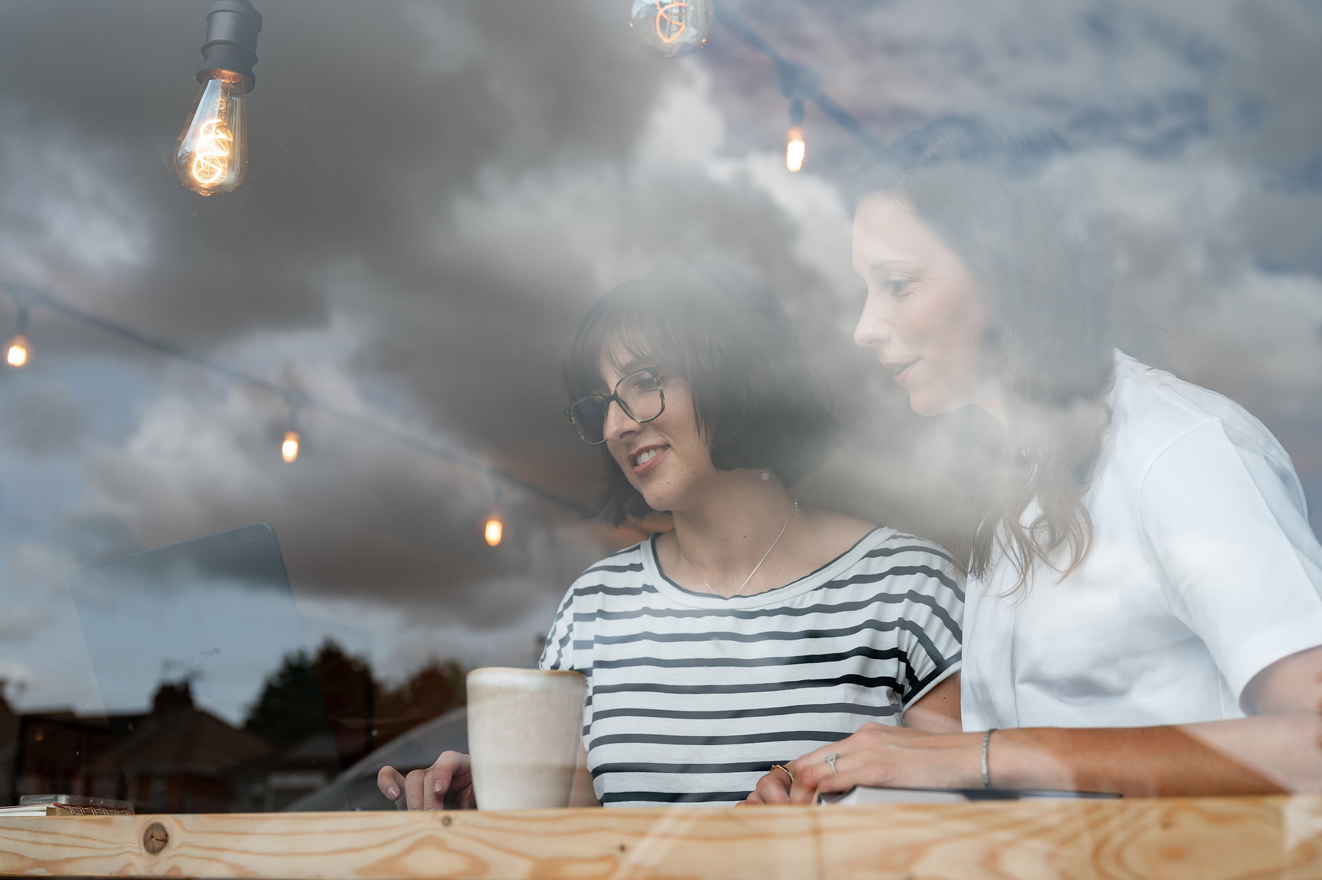 Dr. Becky Cox and Dr. Chelcie Jewitt from Surviving in Scrubs sit indoors at a wooden table, engaged in a brand shoot, as they work on a laptop with a coffee mug nearby. Reflections of clouds and string lights dance on the glass.