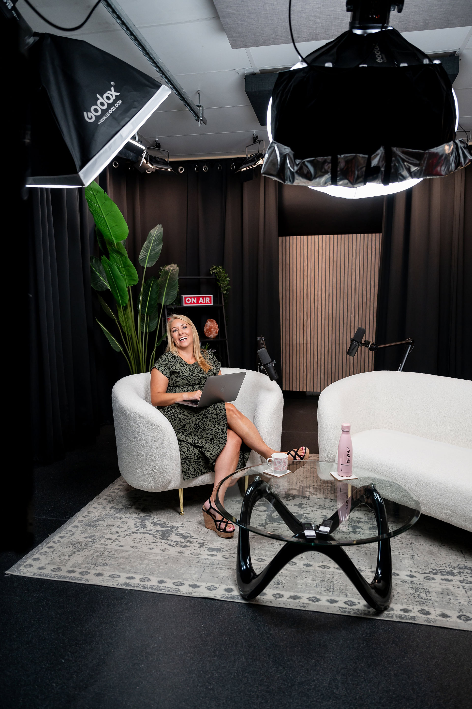 Woman seated on a white couch in a studio with professional lighting. She is holding a laptop and smiling. A "ON AIR" sign, microphone, and glass table with a water bottle are visible.
