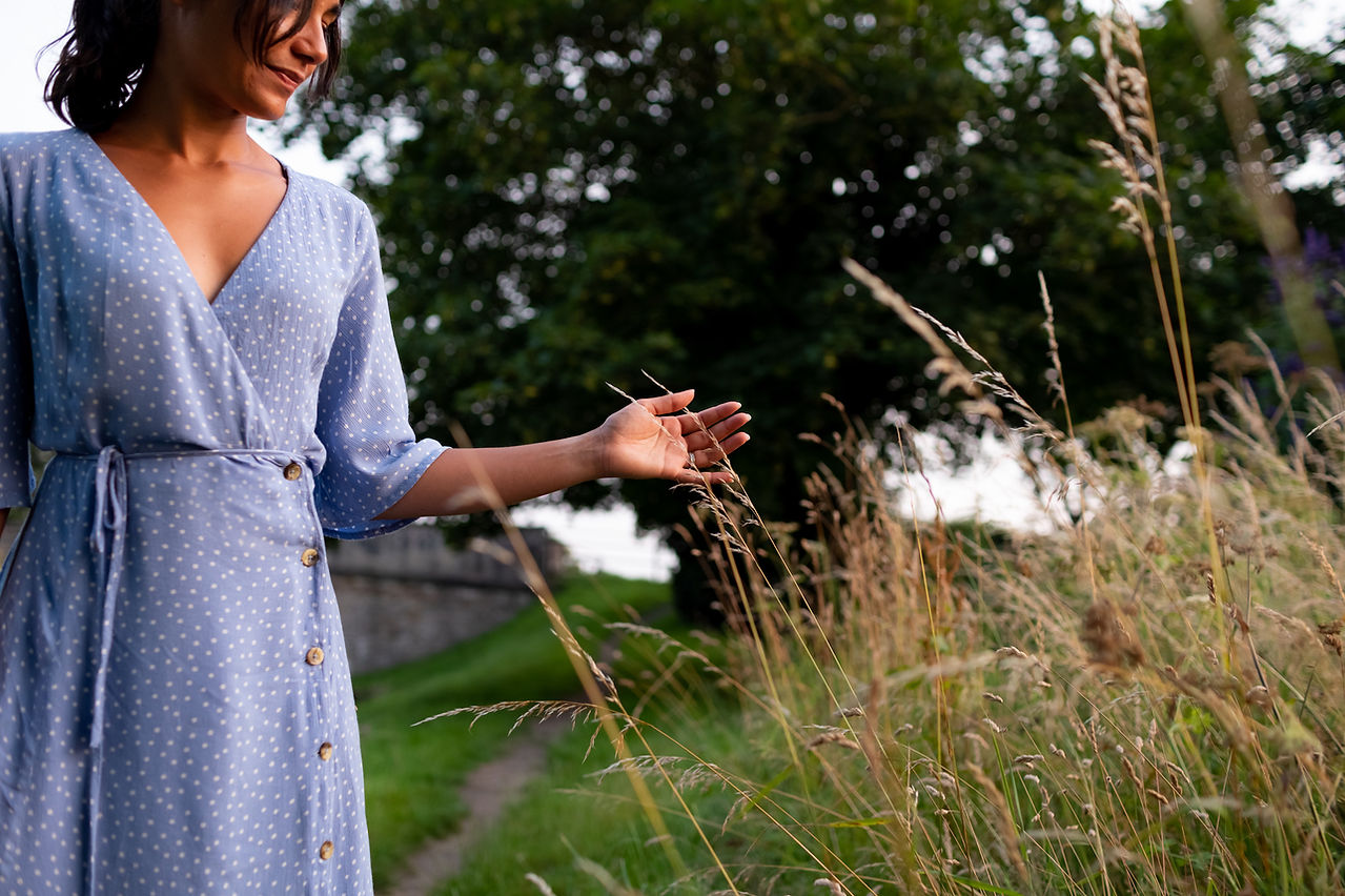 A person in a blue dress with white polka dots touches tall grass in a field, with trees and a path in the background. Captured by a personal brand photographer, this serene scene is perfect for Liverpool brand photography and entrepreneur portraits.