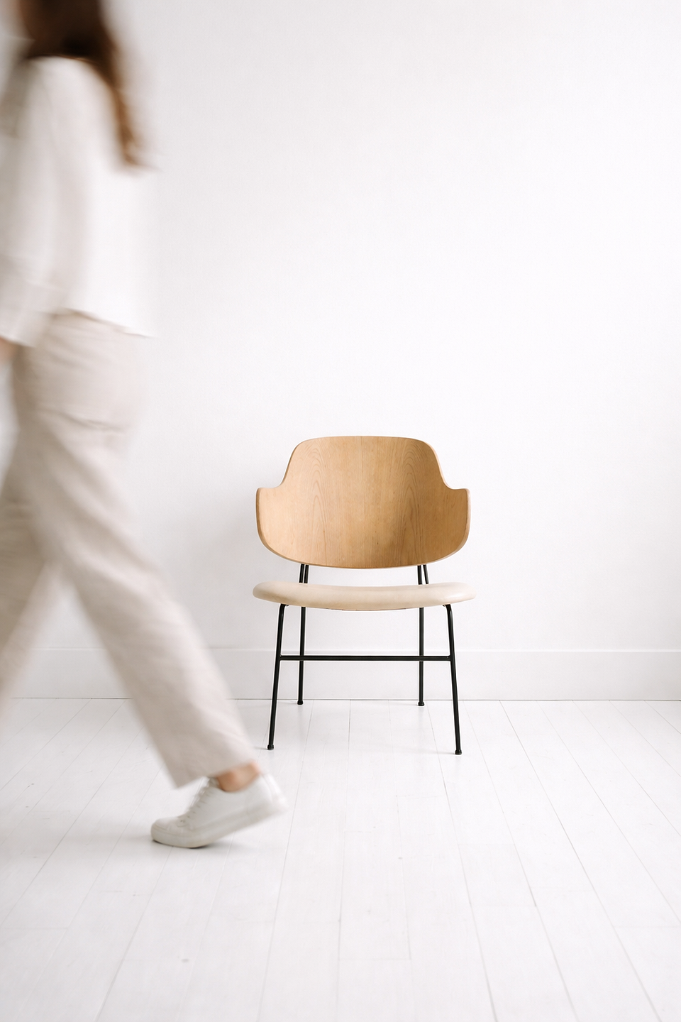 A blurred figure walks past an empty modern wooden chair in a white minimalist room, evoking stillness or absence.