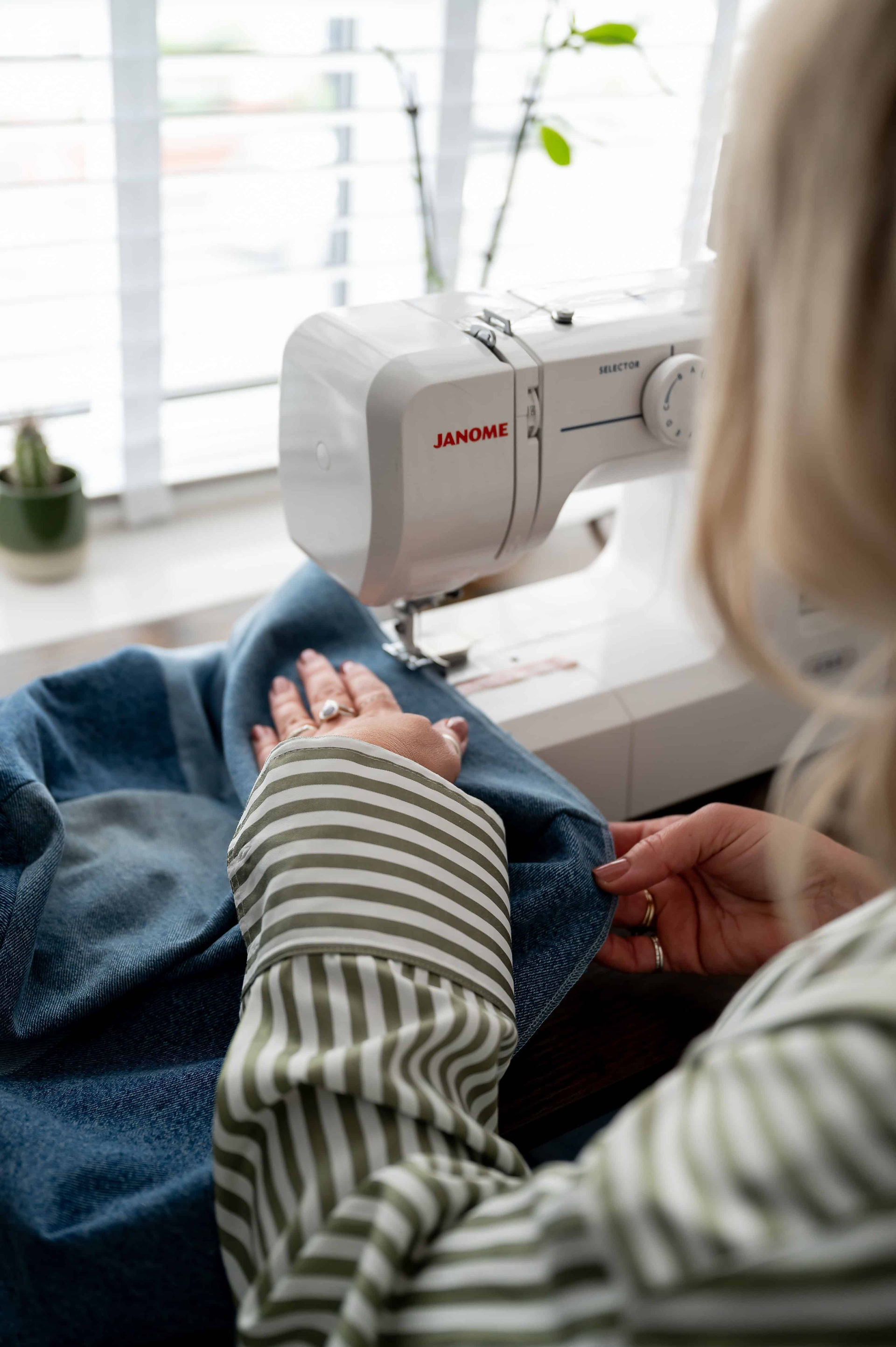 A person is using a Janome sewing machine to sew a piece of blue fabric, with a plant visible in the background. Captured by a personal brand photographer, the scene emphasizes creativity and the serene workspace.