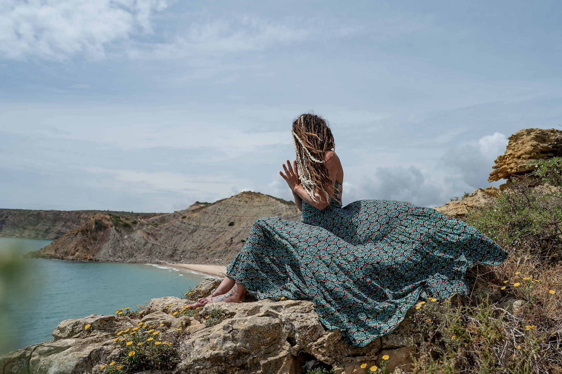 Yoga instructor sat on Algarve cliff in flowing dress in prayer pose with dreadlocks blocking her face.