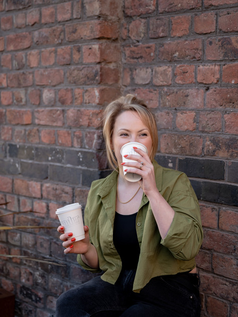 Rachael Lyons sits outdoors against a brick wall, smiling and sipping coffee from a white takeaway cup, wearing a green shirt and black top.