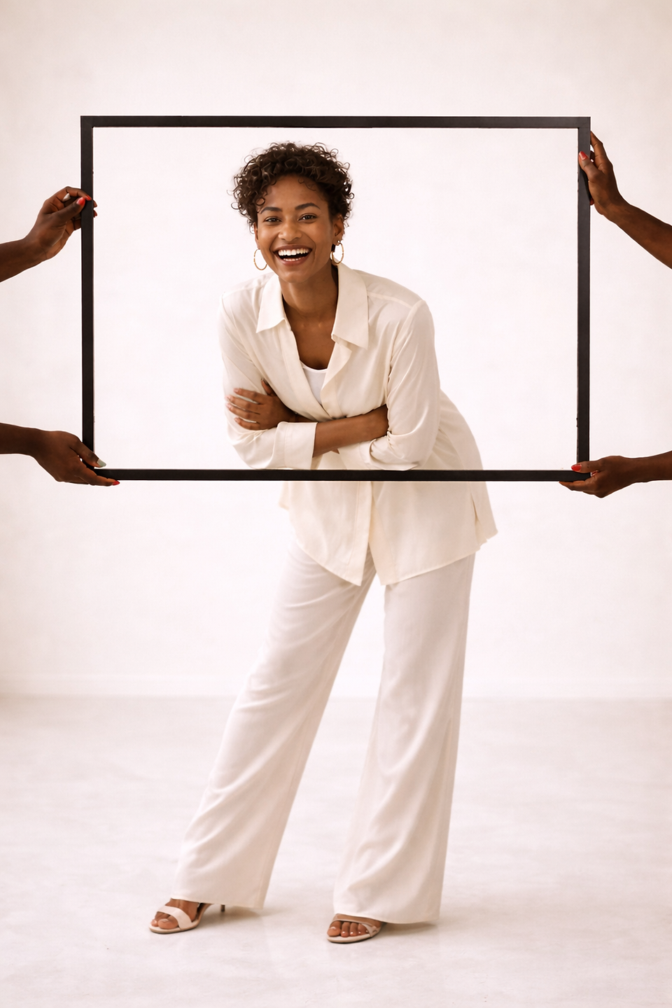 A smiling woman dressed in an off-white set is framed within a rectangular black picture frame held by multiple hands, evoking visibility, perspective, and empowerment.