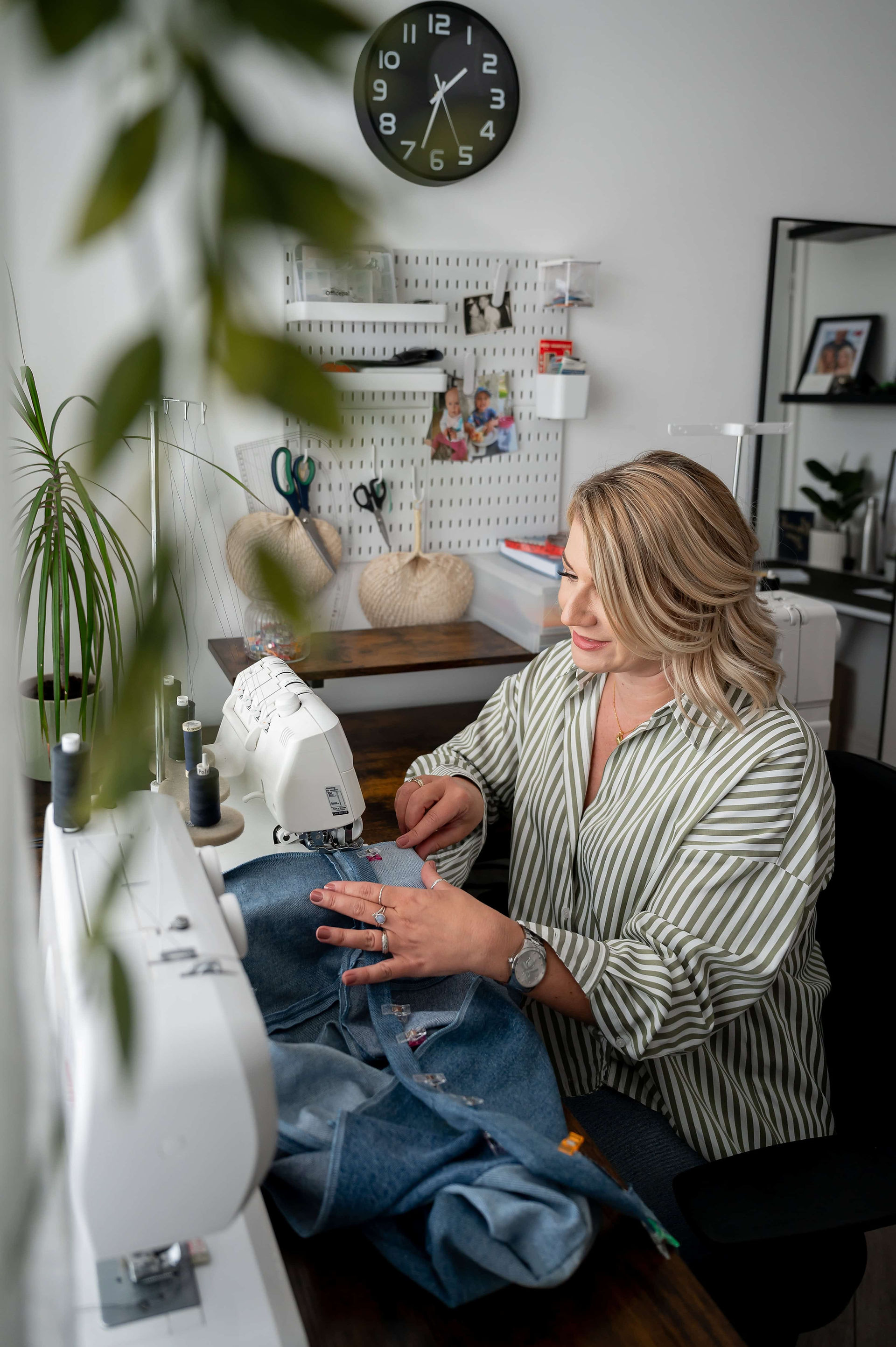 A woman sits at a sewing machine, working on a piece of denim fabric in a well-organized room with various tools and supplies on a pegboard in the background, captured by a UK branding photographer.