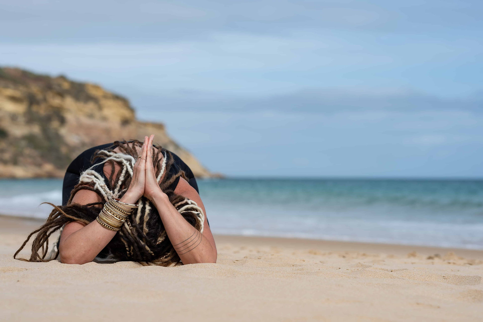 Yoga instructor in childs pose on Burgau Beach in Portugal with sea and cliffs in background.
