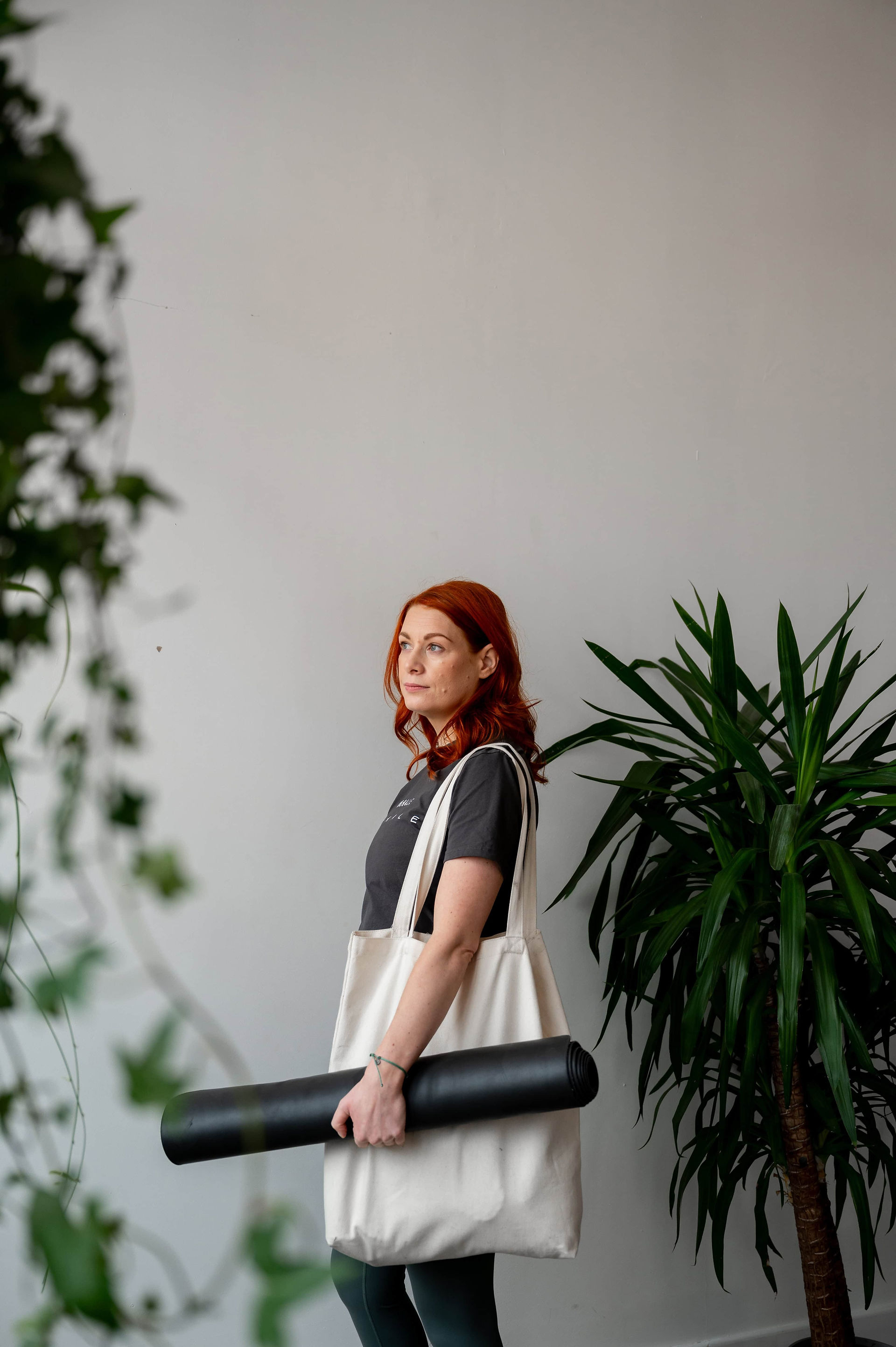 A person with red hair stands against a white wall, carrying a rolled-up yoga mat and a white tote bag, with indoor plants in the foreground and background. Captured by Wild Kind Photography, this image beautifully blends lifestyle and serenity.
