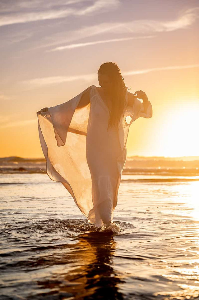 A woman in a flowing white dress walks through shallow sea water at sunset, holding up her dress for a brand photoshoot, backlit by the golden sunset in Portugal..