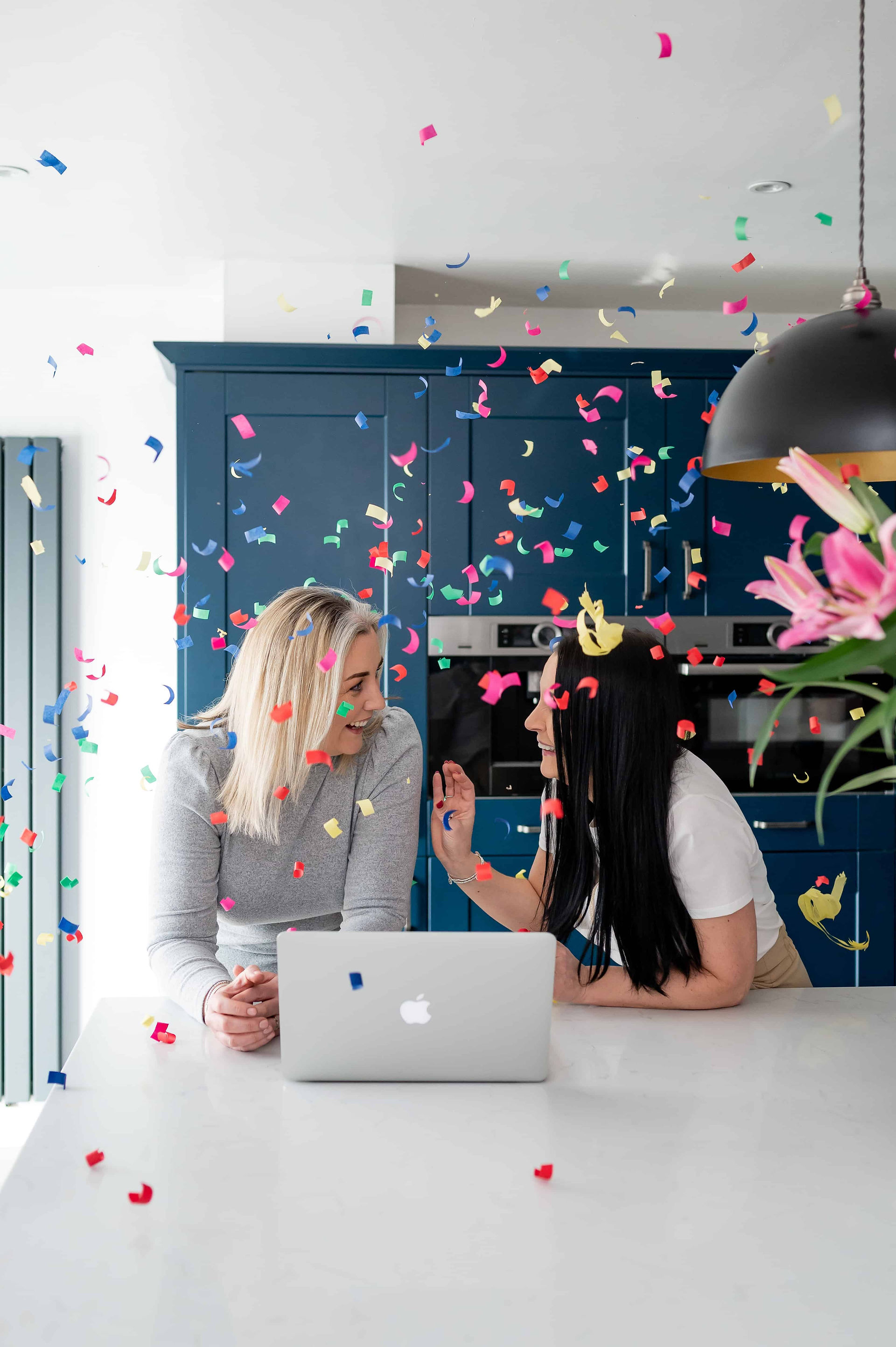 Podcast hosts laughing while colorful confetti rains down in kitchen.