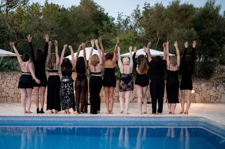 Group of women in black dresses and lingerie holding hands in the air, facing away from the camera by the pool in Mallorca.