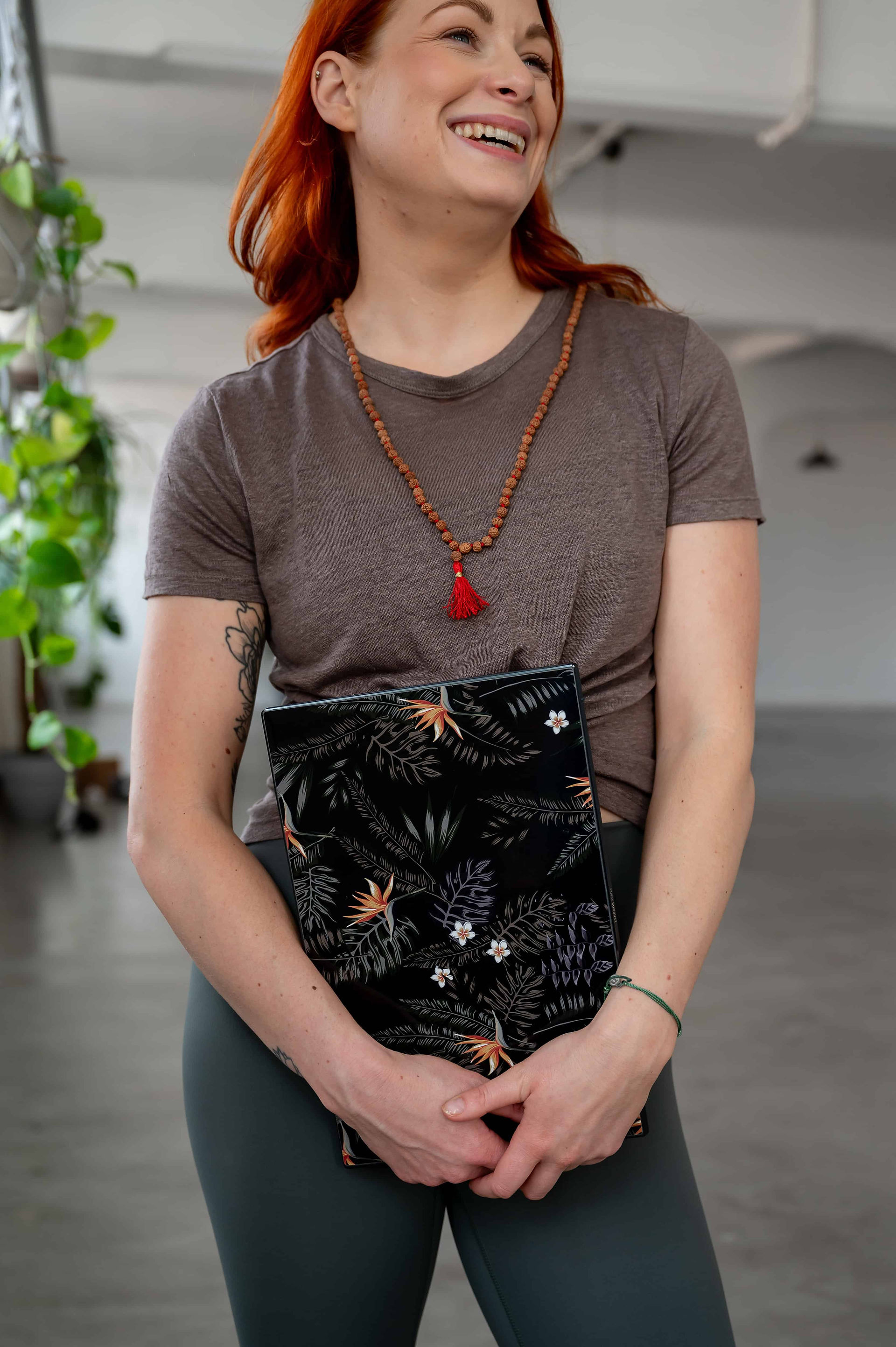 A person smiles while holding a floral-patterned book. They wear a brown t-shirt, green pants, and a beaded necklace with a red tassel. Green plants are visible in the background, captured by Wild Kind Photography.
