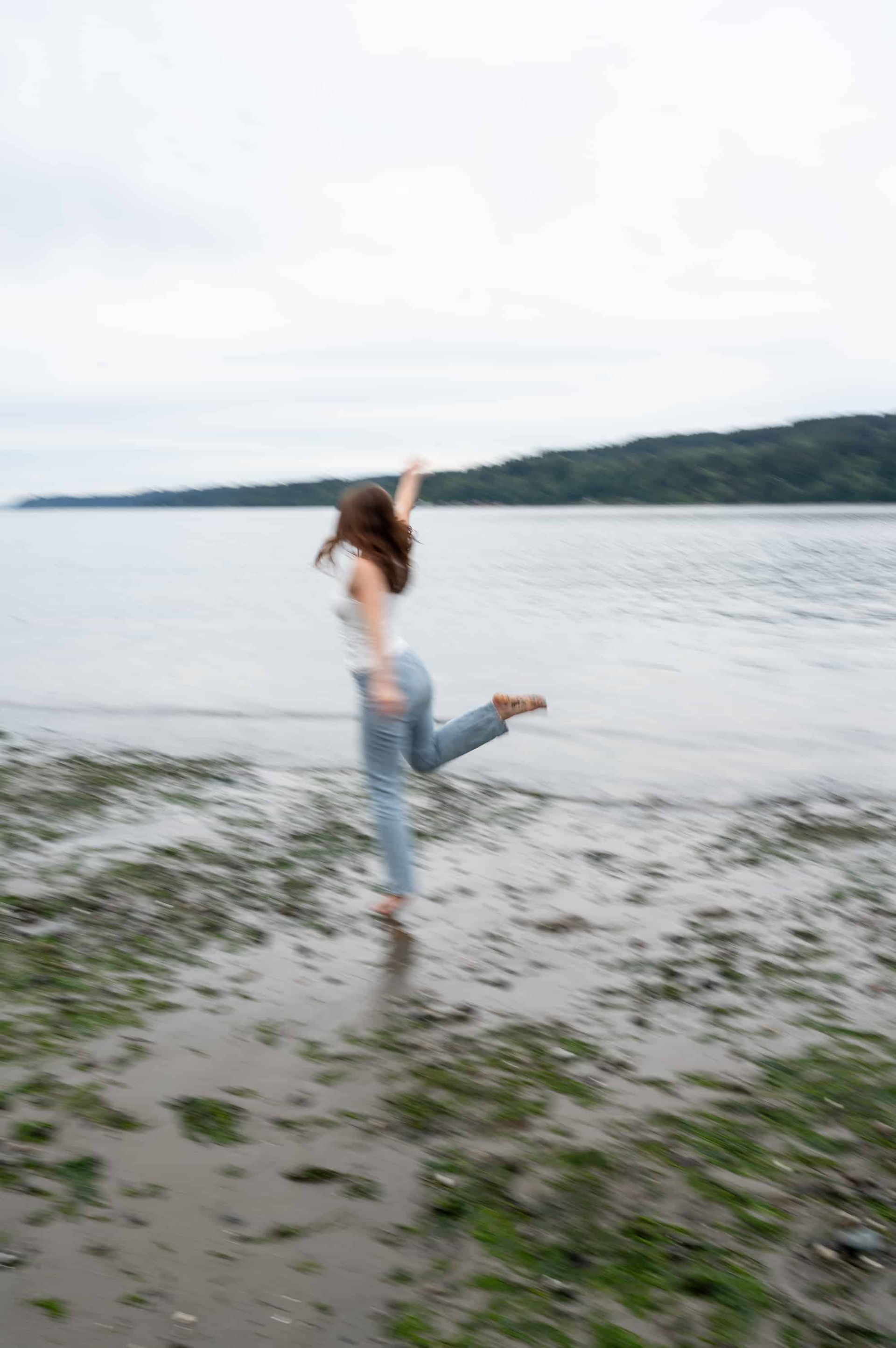 A person in casual clothing is captured in motion by a wild kind photography session, appearing to kick up their leg on a sandy shore, with water and distant greenery in the background.