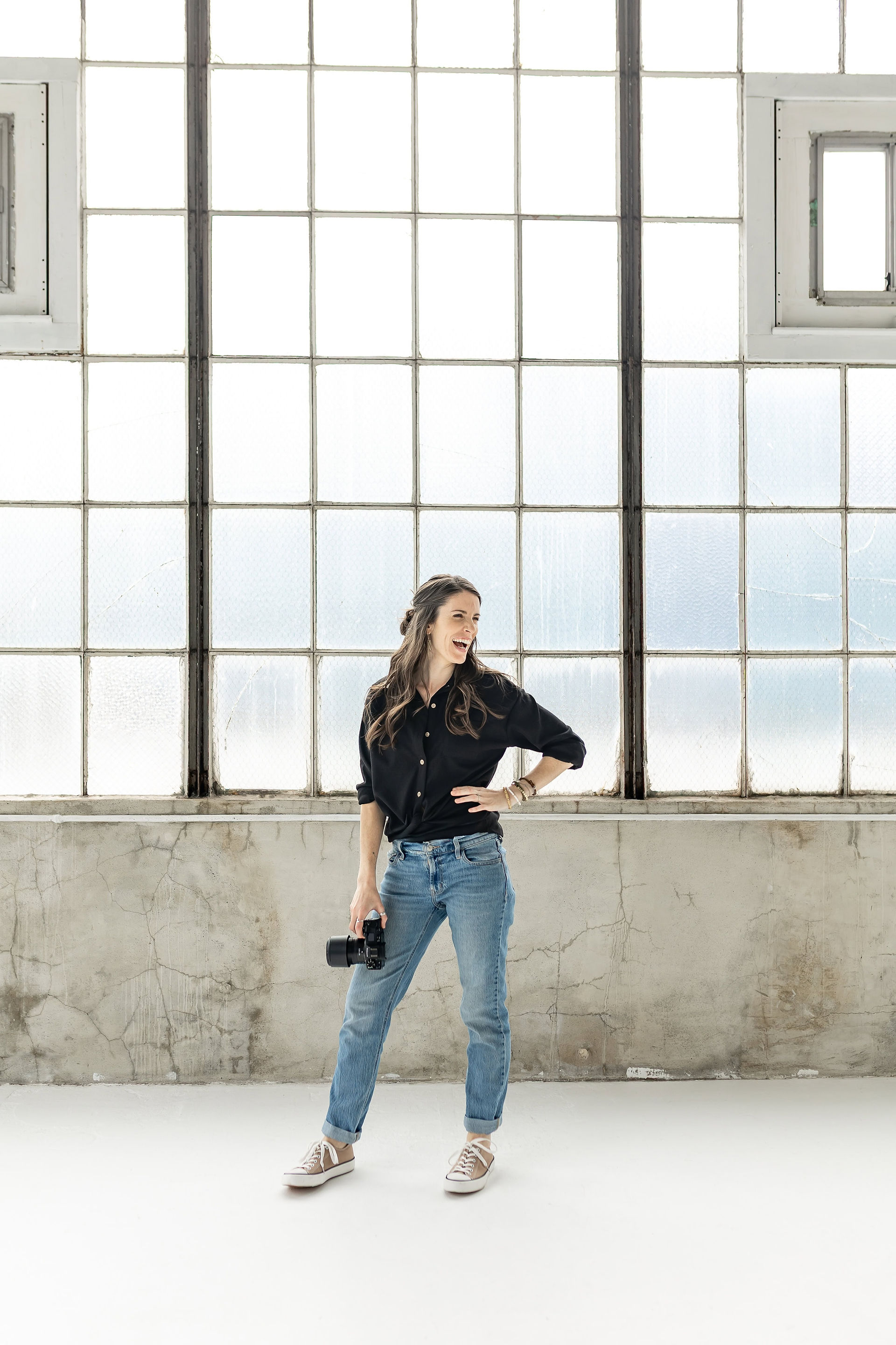 Brand photographer standing with camera smiling sideways with big windows in the background.
