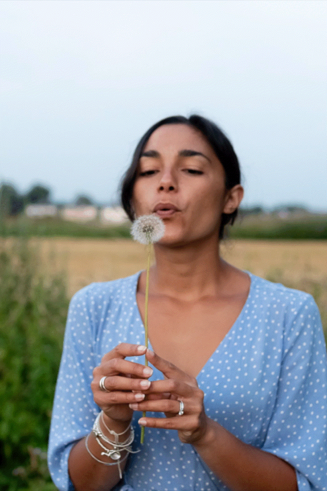 A woman in a blue polka dot dress blows on a dandelion seed head in an outdoor setting with fields in the background, capturing the essence of wild kind photography.
