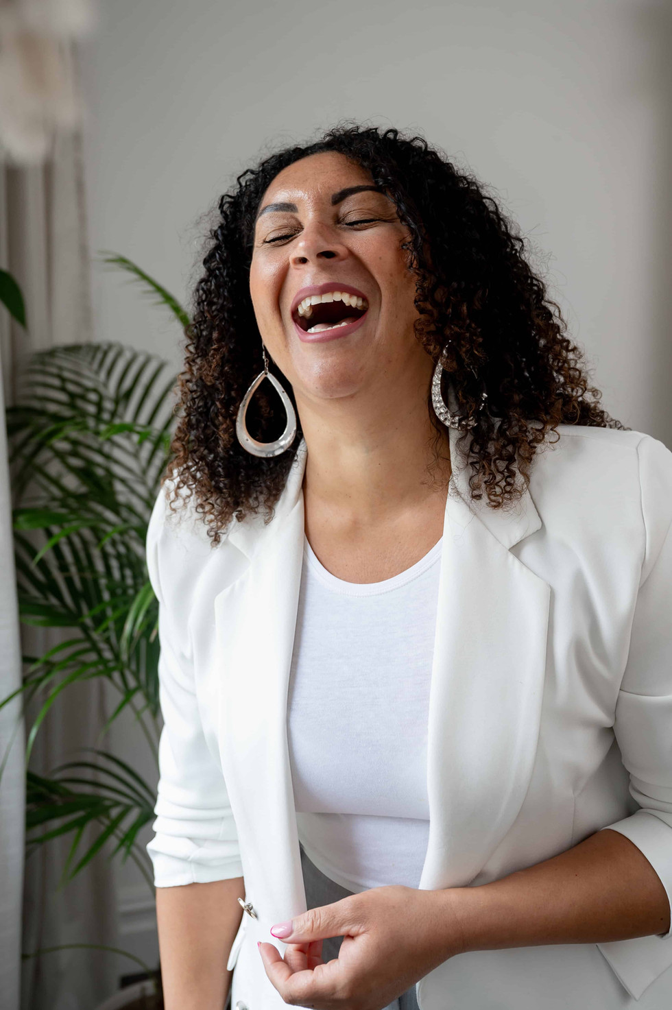Woman in a white blazer laughing joyfully against a minimalist, plant-filled backdrop in a Wirral photo studio.
