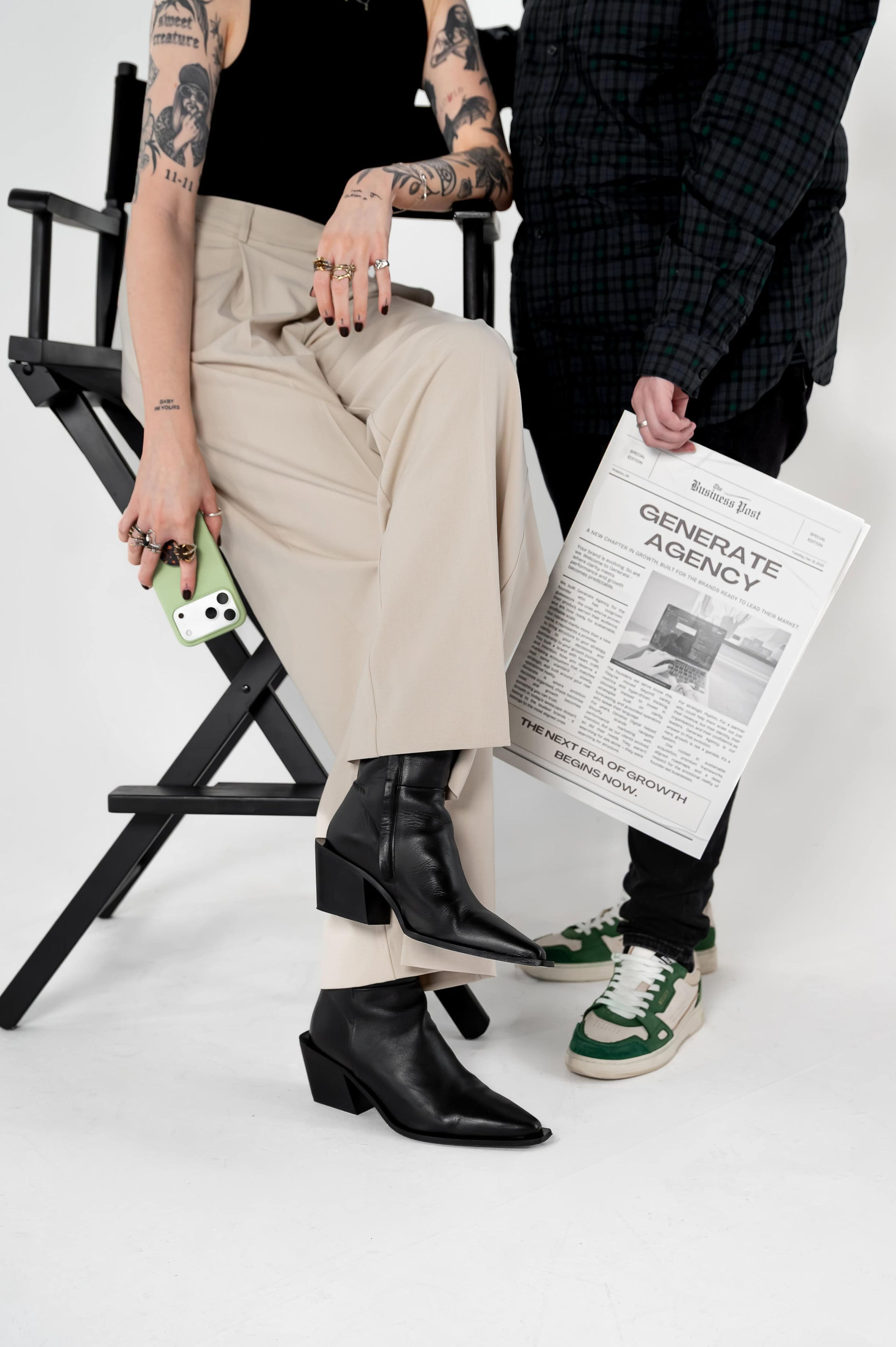 Two business owners posing with creative props during a brand photoshoot, sitting in a director's chair holding a branded newspaper and phone.