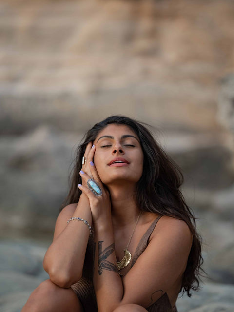 Powerful brand portrait of a feminine embodiment coach meditating with eyes closed, hands on face, at a rocky beach in Mallorca.