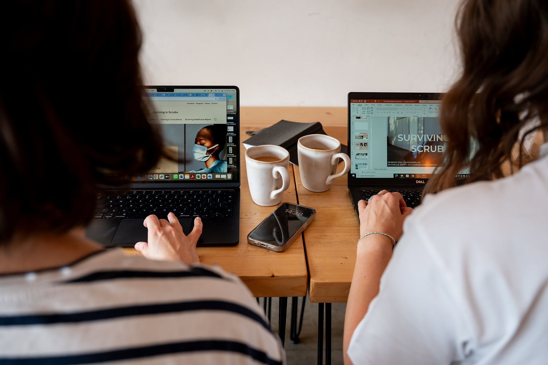 Drs. Becky Cox and Chelcie Jewitt work on laptops at a wooden table, surrounded by coffee mugs and a smartphone during their brand shoot. One screen displays a presentation titled "Surviving in Scrubs."