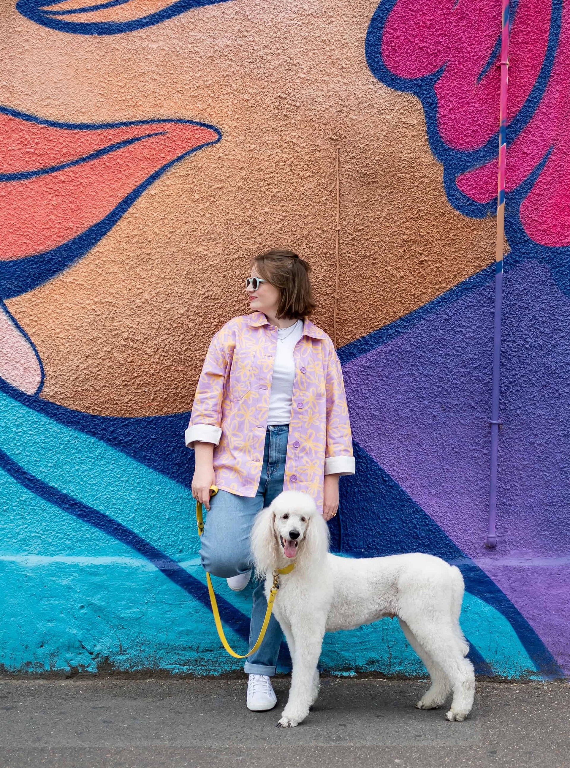 Person standing with a white poodle on a leash in front of a colorful mural on a brand photography shoot.