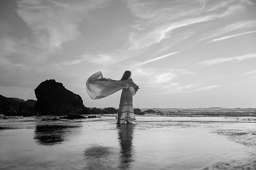 Woman at Portuguese beach looking out to sea with dress blowing in wind at sunset.