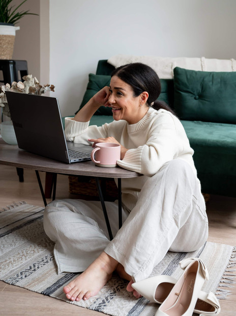 Woman sitting on the floor during a brand photoshoot, leaning on a coffee table with a laptop, laughing naturally in a cozy setting.