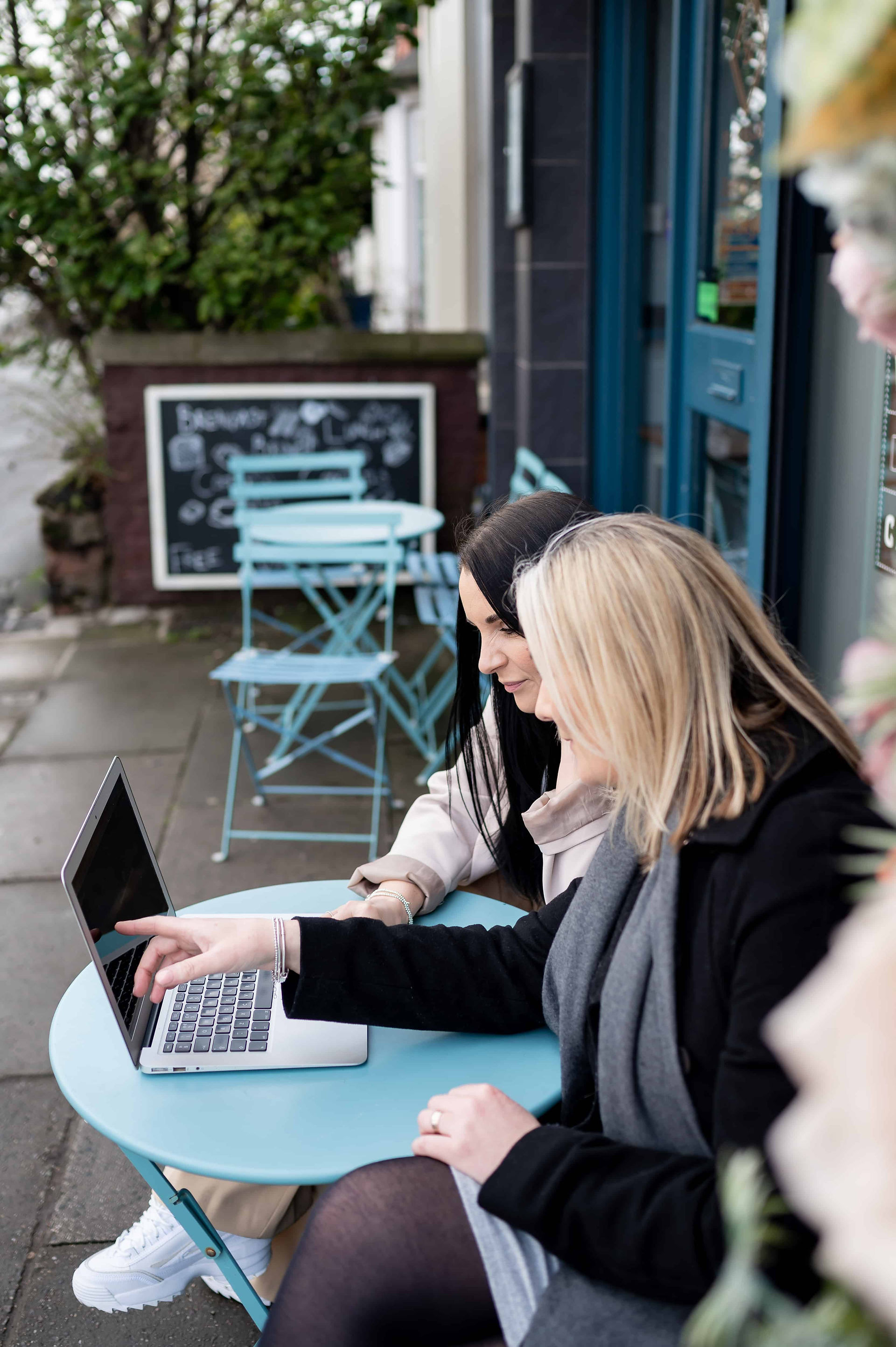 Two people sit at a blue outdoor table, looking at a laptop. One points at the screen. A chalkboard and more chairs are in the background.