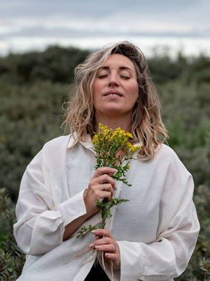 Blonde woman in a white shirt holding yellow flowers and smiling with eyes closed in an open field. Wild and intuitive branding session surrounded by greenery.