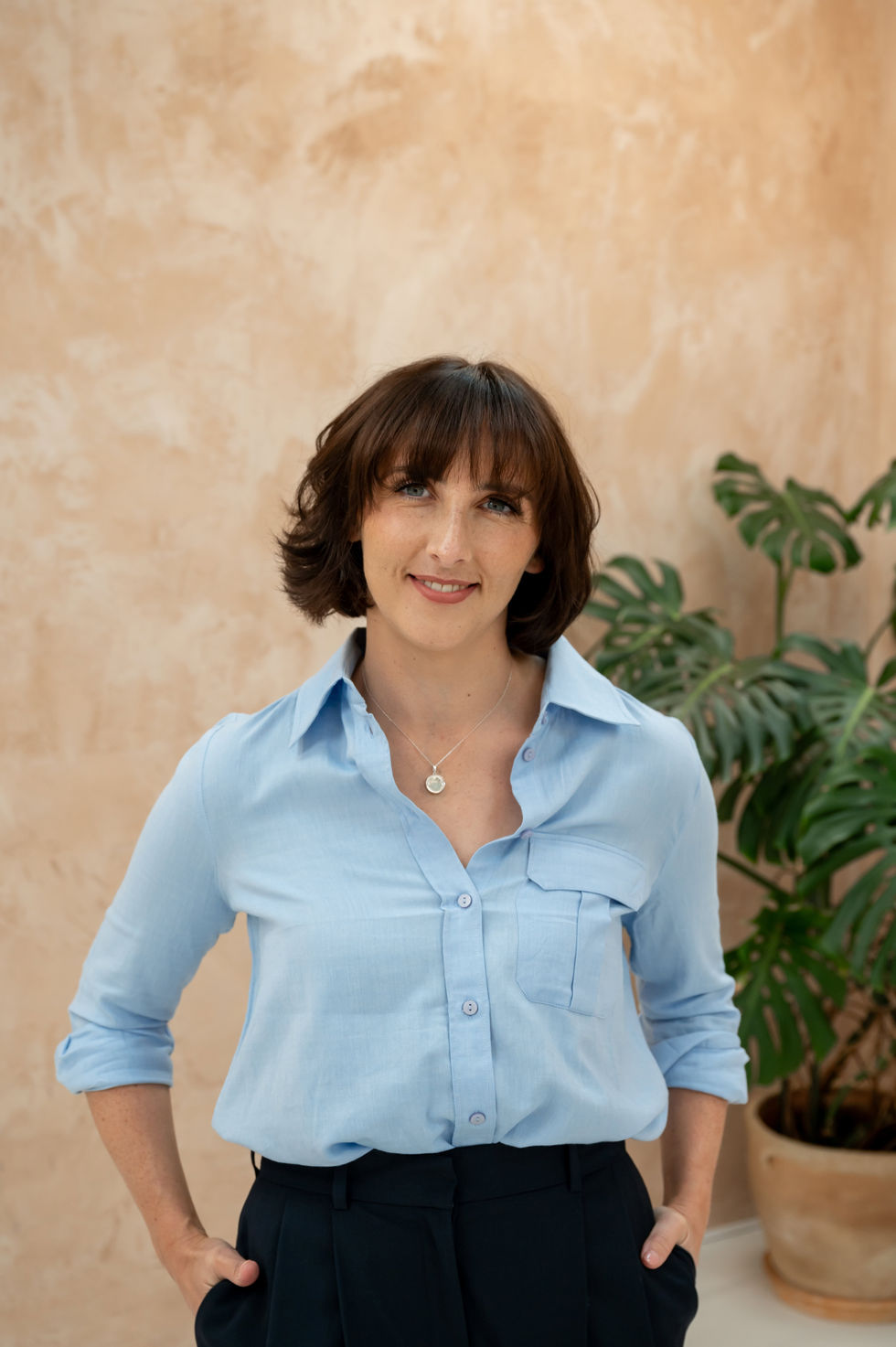 A woman with short brown hair in a light blue shirt stands in front of a beige wall adorned with a lush green plant, embodying elegance and professionalism during a brand shoot.